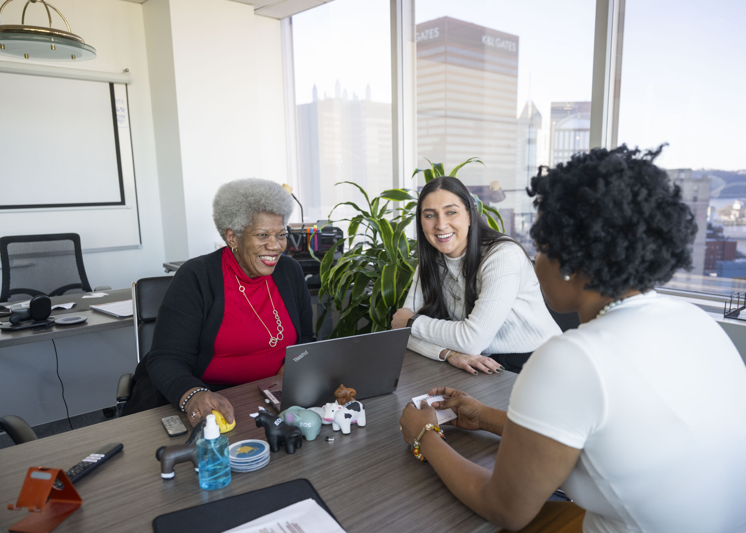An older Black woman, a younger Black woman and a younger white woman sit and laugh together at a conference table in an office in Pittsburgh, Pennsylvania.