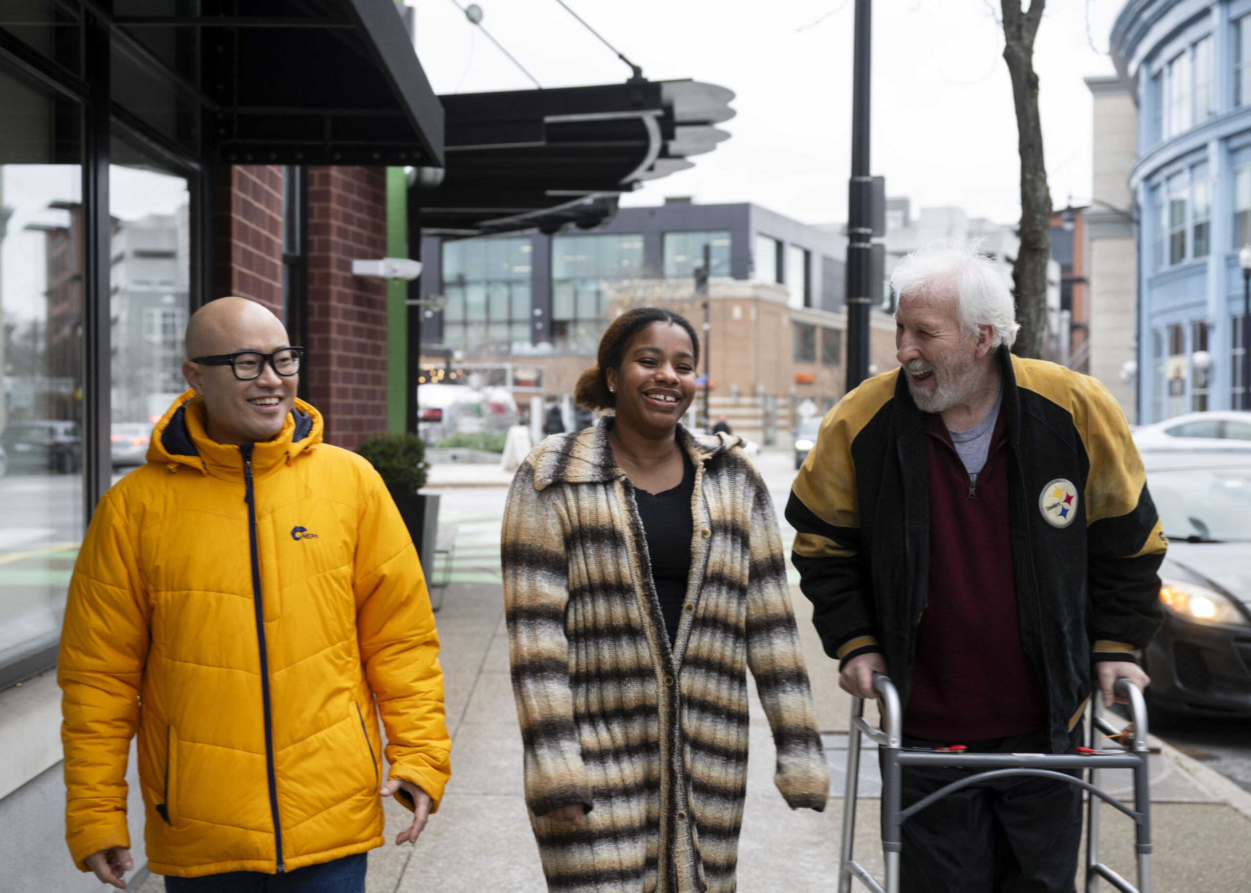 An older white man is wearing a Pittsburgh Steelers jacket and using a walker at SouthSide Works in Pittsburgh, Pennsylvania. He is walking and talking with a younger Black woman and a younger Asian man in a bright yellow jacket.