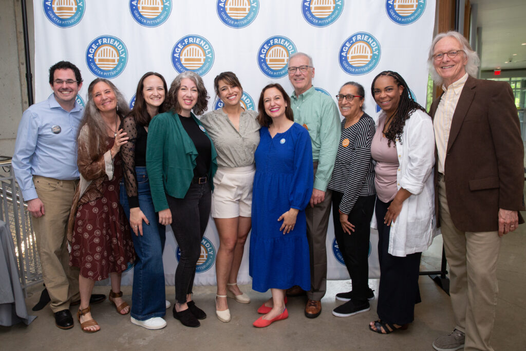 Ten people of all races, genders and ages pose for a group picture in front of the Age-Friendly Greater Pittsburgh logo.